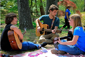 Three teens sit on a picnic blanket playing each playing a guitar and smiling together.