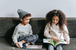 A young boy and girl sit on the couch reading books together.