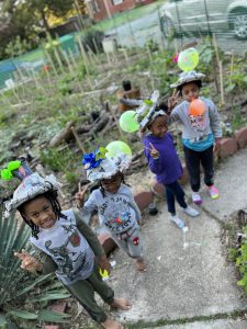 Four children wearing newspaper hats of flowers stand in a garden. 