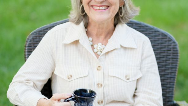 Julie Bogart sitting in a black wicker chair, in a white suit, with a cup of coffee smiling warmly at the camera